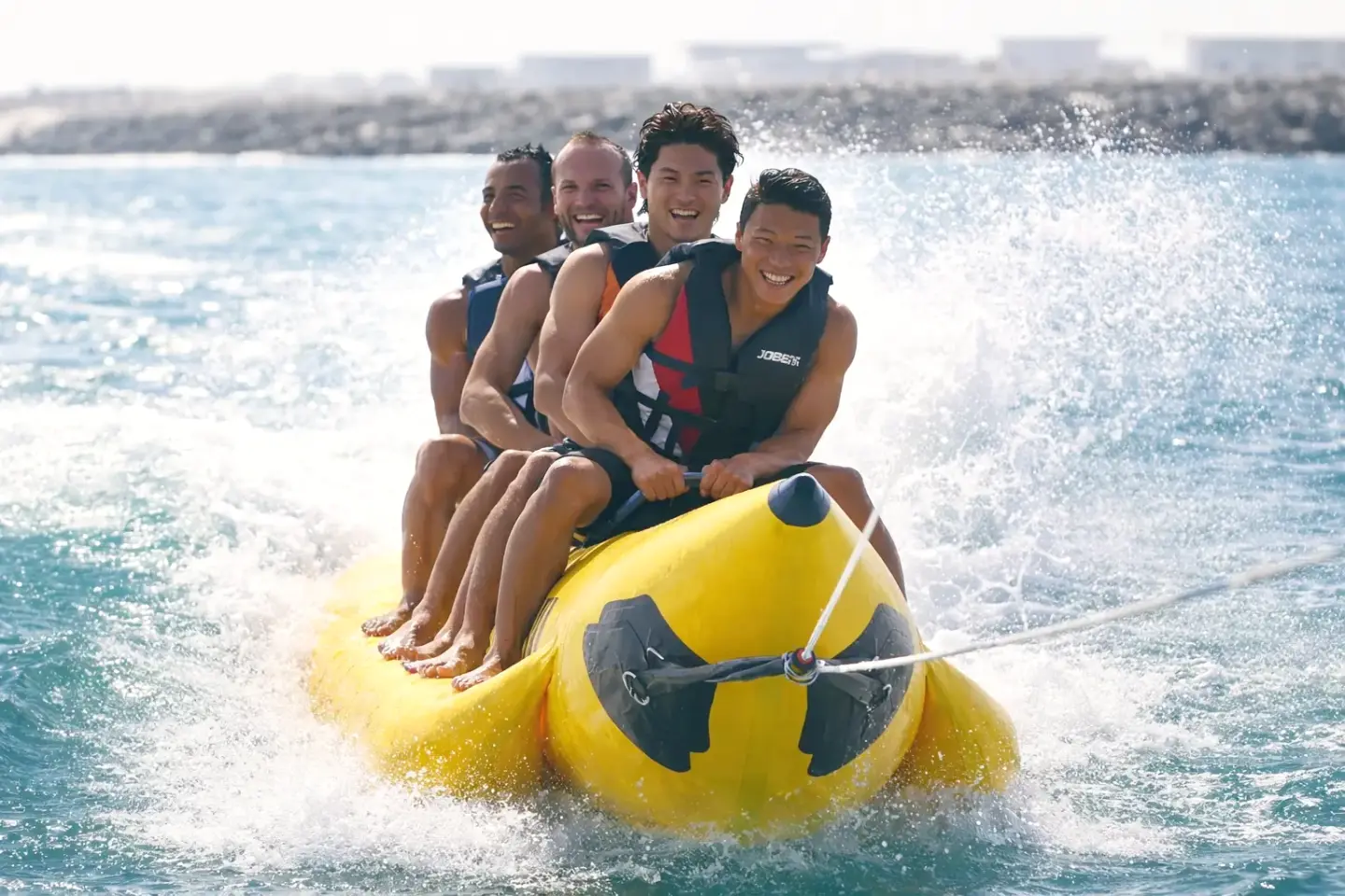 Four smiling riders in life vests holding on to a yellow banana boat through spray
